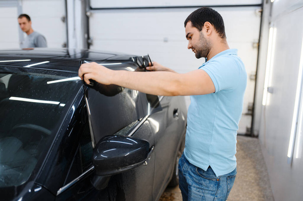 男性労働者は濡れ車の着色、チューニングサービスをしようとします。ガレージの車窓にビニールの色合いを適用する機械、着色された自動車用ガラス - 写真・画像