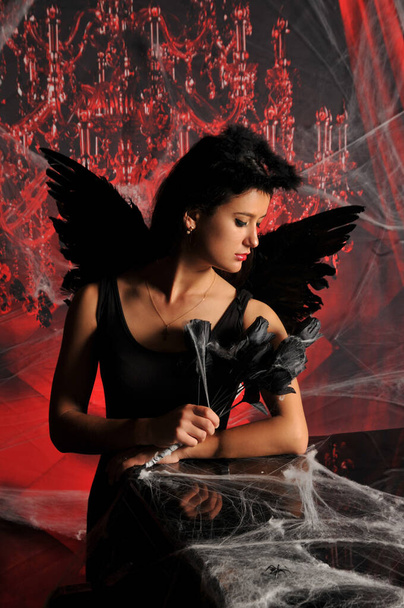 Beautiful young brunette woman in black angel costume with wings over spooky red background posing by piano with rose bouquet. Halloween party outfit - Photo, Image