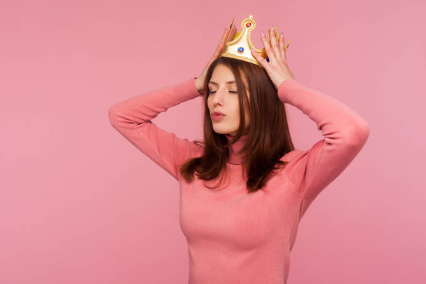 Selfish confident brunette woman adjusting golden crown on her head, proud of her status, pretending to be queen. Indoor studio shot isolated on pink background - Valokuva, kuva