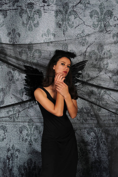 Beautiful young brunette woman in black angel costume with wings over dark spooky background. Halloween party outfit - Foto, Imagem