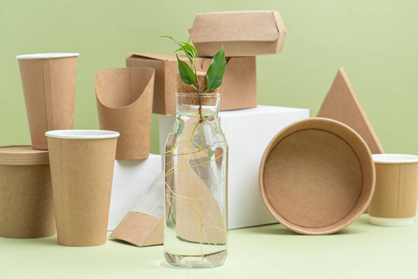 Eco-friendly disposable paper containers for food and drinks over green background. Side view on composition. Some objects placed on white cube pedestals, some placed on side. Plant branch in bottle. - Foto, Imagem