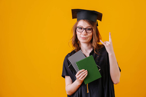 Young woman in glasses celebrating her graduation. She's wearing gown and hat. Over orange background. Posing for camera, making rock n roll gesture. - Valokuva, kuva