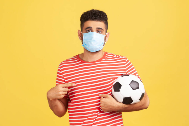 Young bearded man in safety mask and red t-shirt pointing finger at soccer ball on his hand with sad expression, coronavirus quarantine. Indoor studio shot isolated on yellow background - Photo, image