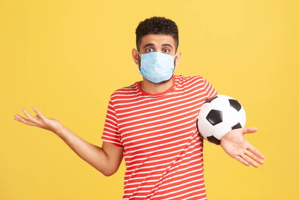Frustrated uncertain man in red t-shirt and hygienic face mask spreading hands holding ball, confused how to play football safety. Indoor studio shot isolated on yellow background - Photo, image