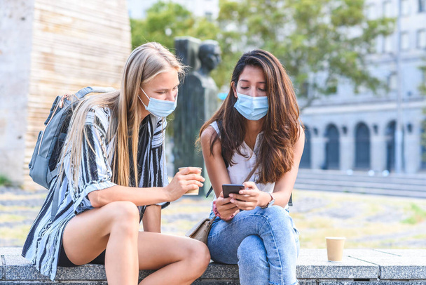 dos jóvenes hermosas amigas sentadas en un banco de piedra en la calle y con máscaras higiénicas azules que se muestran la pantalla del teléfono inteligente - Foto, Imagen