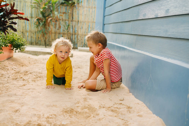 Outdoor summer portrait of two cute happy playing kids, big brother and his little sister against blue house wall. Girl is on all fours and boy is looking at her. - Foto, Imagen