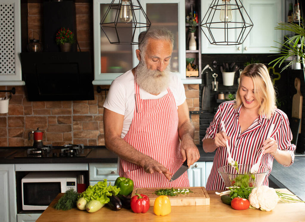 Senior couple having fun in kitchen with healthy food. Man and woman preparing lunch with bio vegetables. Happy elderly concept with mature funny pensioner. - Foto, Imagen