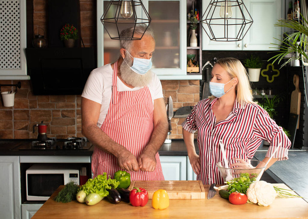 Happy caucasian aged couple cooking at home in face mask. Making salad at home. Home cooking. Social distance. Cooking during covid 19. - Fotó, kép