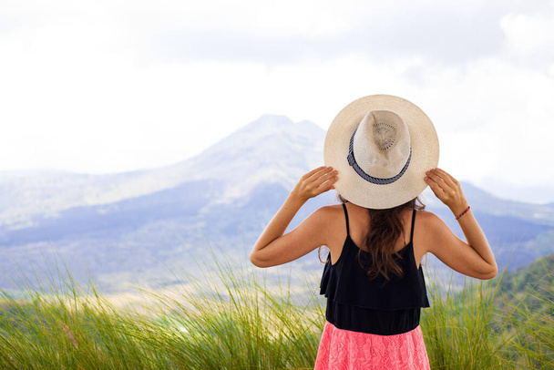 Young traveler woman wearing hat, enjoying mountain landscape. Hills and Batur volcano. Scenic panoramic view. Sky with white clouds. View from back. Copy space. Kintamani, Bali, Indonesia - Foto, imagen