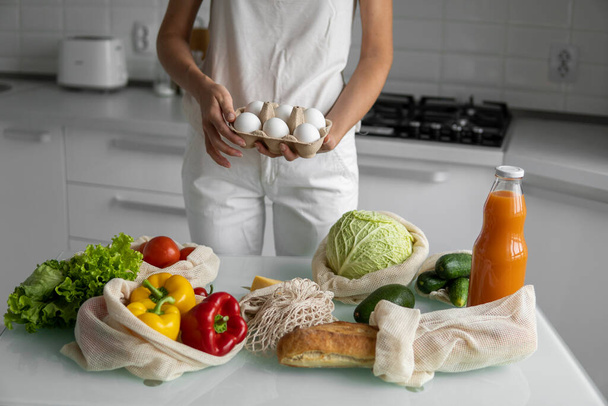 Woman holding eggs tray with a reusable shopping bags with vegetables on a table on a kitchen at home. Zero waste and plastic free concept. Mesh cotton shopper with vegetables. Ecology. - Fotoğraf, Görsel