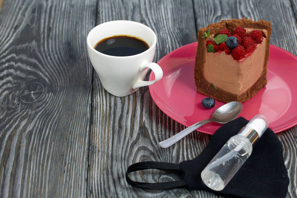 A piece of no-bake chocolate cake with cream cheese. Garnished with raspberries, blueberries and mint leaves. Next to it is a cup of coffee and a medical mask with antiseptic. - Φωτογραφία, εικόνα