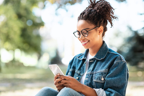 Μαύρο κορίτσι με Smartphone χρησιμοποιώντας το κινητό App κάθεται στο πάρκο - Φωτογραφία, εικόνα