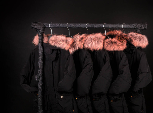 A lot of black winter coats, jacket with fur on hood hanging on clothes rack on black background, black friday - Photo, Image