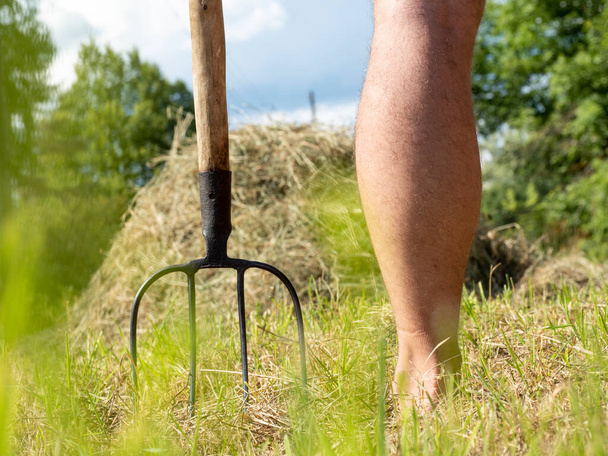 Falusi vasvilla egy férfi lába mellett. A háttérben egy szénakazal. A mezőgazdaság fogalma. Napsütéses nyári nap - Fotó, kép