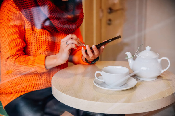 Kafede telefonlu bir kız. Kız elinde bir fincan çayla bir kafede haber bülteni izliyor. Pencereden bak. - Fotoğraf, Görsel