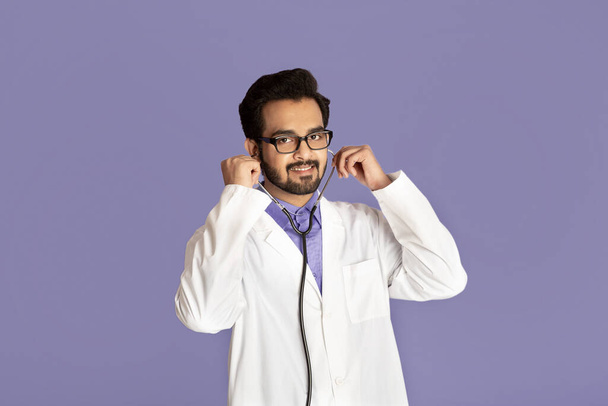 Πορτρέτο του χαρούμενου Ινδού γιατρού σε εργαστηριακή ποδιά με στηθοσκόπιο σε βιολετί φόντο - Φωτογραφία, εικόνα