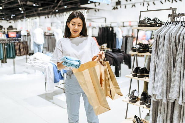 Vrolijke vrouw doet kleren in een tas, kledingwinkel. Vrouwelijke persoon winkelen in de mode boetiek, shopaholic, shopper op zoek naar kleding - Foto, afbeelding