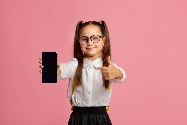 Gadgets for study. Schoolgirl in glasses, school uniform shows a finger at blank screen of smartphone - Photo, Image