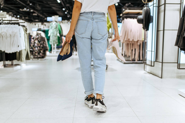 Young woman holds many shoes in clothing store, back view. Female person shopping in fashion boutique, shopaholic, shopper looking on boots - 写真・画像
