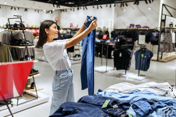 Веселая женщина выбирает джинсы в магазине одежды. Женщина делает покупки в модном бутике, шопоголик, покупатель смотрит на одежду - Фото, изображение
