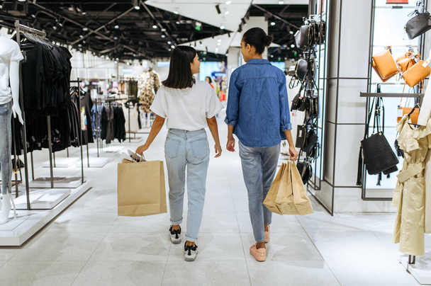 Χαμογελώντας φιλενάδες με χαρτοσακούλες περπατά στο κατάστημα ρούχων. Γυναίκες ψώνια στην μπουτίκ μόδας, shopaholics, αγοραστές με αγορά - Φωτογραφία, εικόνα