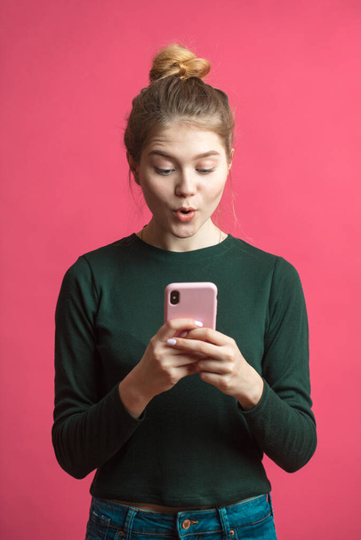Portrait if a shocked young girl with mobile phone isolated over pink background - Foto, immagini