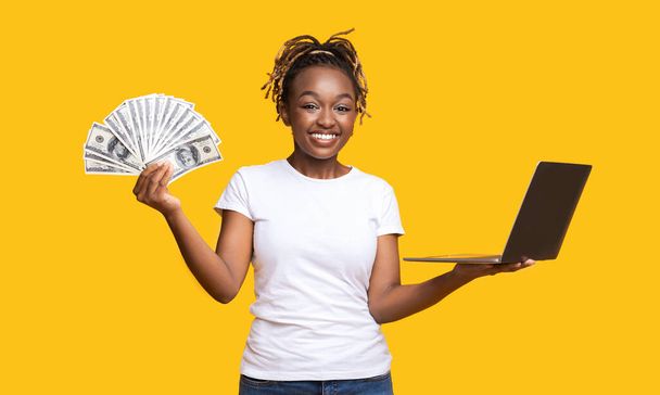 Chica alegre negro con dinero en efectivo y portátil en amarillo - Foto, Imagen