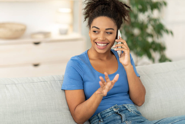 Šťastná africká dáma mluví na mobilní telefon sedí na gauči uvnitř - Fotografie, Obrázek