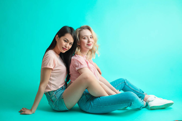 two multiethnic women sitting on the floor back to back isolated on blue background - Фото, зображення