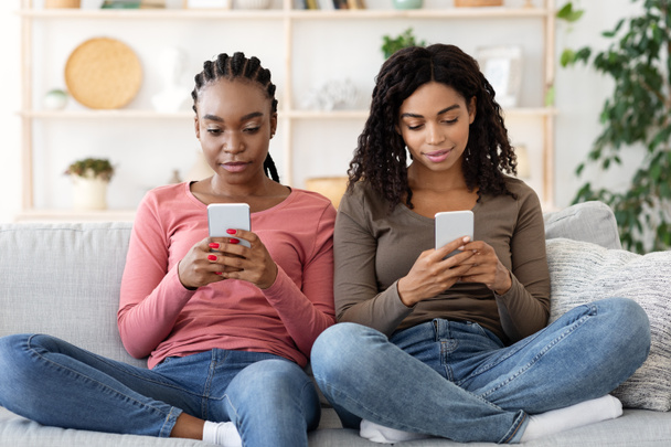 Zwarte meisjes met mobiele telefoons zitten op de bank thuis - Foto, afbeelding
