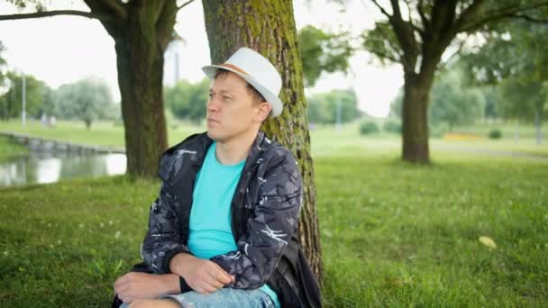 Atraktivní muž v klobouku odpočívající pod stromem v městském parku, sledovací kamera - Záběry, video