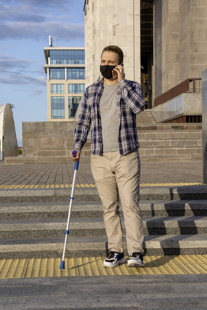 Pouliční portrét nemocného mladého muže v lékařské masce, který chodí po městě s berlí. Možná utrpěl zranění nohy a doktoři mu nařídili, aby šel na čerstvý vzduch.. - Fotografie, Obrázek
