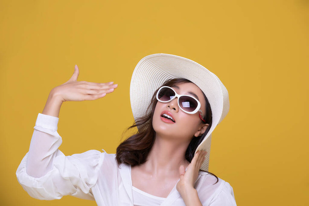 Портрет Азіатської прекрасної щасливої молодої жінки з сонцезахисними окулярами і капелюхом, що радісно посміхається влітку і дивиться на фотоапарат ізольований на жовтому студійному тлі. - Фото, зображення