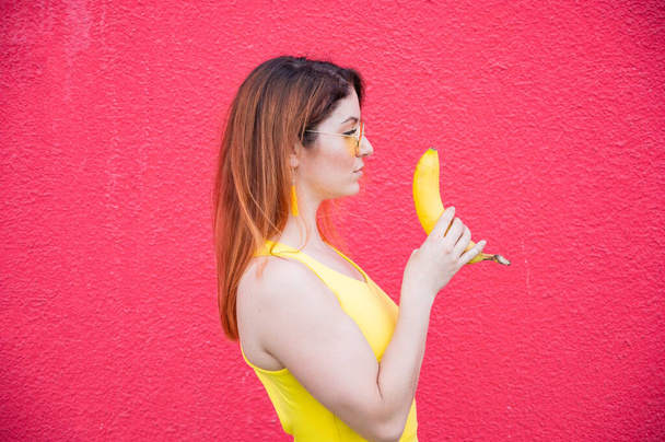 Red-haired woman in sunglasses holds a banana on a red background - Φωτογραφία, εικόνα