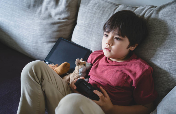 Portrait of kid holding video game or game console. Child playing game online at home, Young boy siting alone on sofa relaxing on his own on weekend, New normal lifestlye concept - Photo, Image