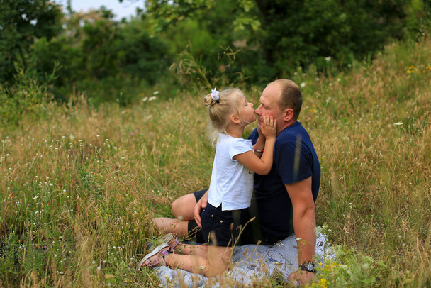 La figlia bionda di tre anni bacia papa 'sul naso, abbracciandogli delicatamente le guance. Papà e figlia sono seduti nell'erba nella natura. L'amore di suo padre per sua figlia. Vacanza in famiglia nella natura. - Foto, immagini