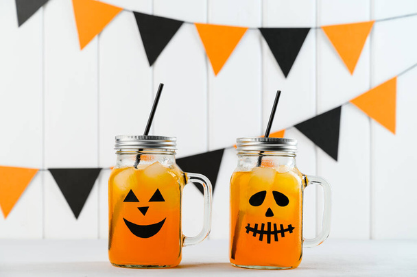 Halloween pumpkin iced mocktails in glass jars decorated with scary faces on a white table. DIY Halloween Party decoration on the wall. - Photo, Image