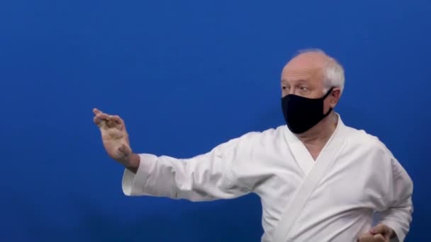 Sobre un fondo azul en una máscara médica negra, un viejo atleta entrena bloques y golpes con un cambio de postura - Metraje, vídeo