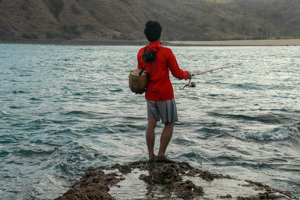 Οι ψαράδες πιάνουν ψάρια με χαμηλή παλίρροια. Θέα από το πίσω μέρος του άντρα με καλάμια ψαρέματος στο νερό. Κοντινό πλάνο του ινδονήσιου άνδρα με ένα καλάθι μπαστούνι στην πλάτη του με κόκκινη κουκούλα πιάνει ψάρια στο καλάμι ψαρέματος - Φωτογραφία, εικόνα
