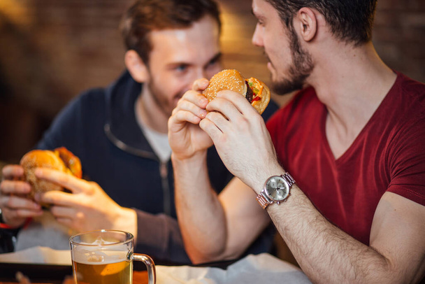 Two happy male friends eating tasty burgers in bar. - Photo, image