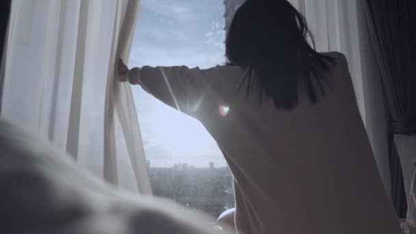 Jonge vrouw wordt wakker en stretching in het bed in de ochtend, open gordijnen, kijken door het raam met uitzicht op de stad straat, hoge verdieping appartement, nieuwe zon met een goede dag  - Video