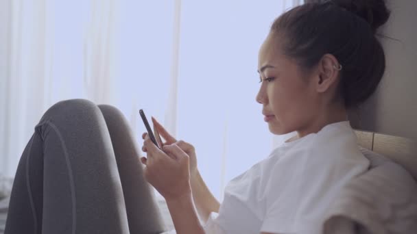 Jovem bronzeado mulher asiática Sente-se de joelhos usando smartphone na cama, conexão de rede on-line, novos feeds ocupado notificações da manhã, confortável janela do quarto luz do dia, vibe de quarto solitário - Filmagem, Vídeo