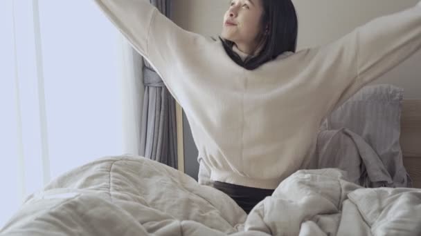 Genç Asyalı kadın kalkar ve kollarını yatağa uzatır, gün doğumunda sabah ışığı, evde rahat yatak odası perdeleri açık, doğal gün ışığı ferahlığı, sıcak kış süveteri giyer. - Video, Çekim