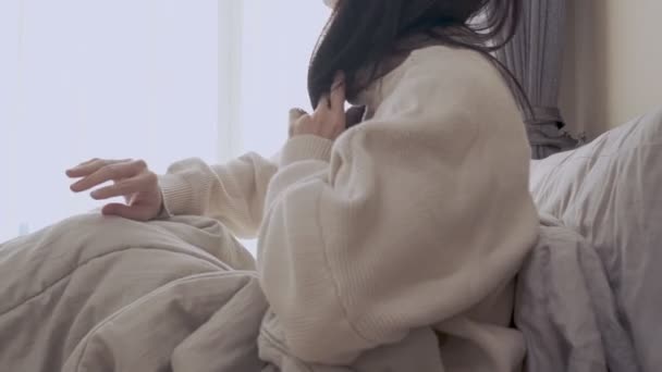 Asian female in sweater wakes up in warm bed morning sunshine inside the bedroom, side view, lazy Day messy blanket bed white curtains, natural day light sun lens flare, luxury interior design bedroom - Footage, Video
