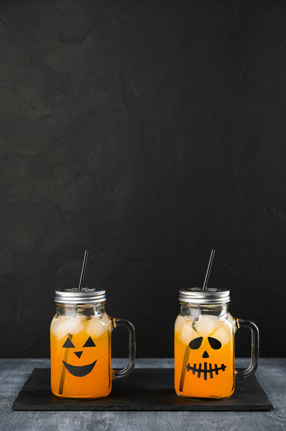 Halloween pumpkin mocktails in glass jars decorated with scary faces on the black background. - Photo, Image