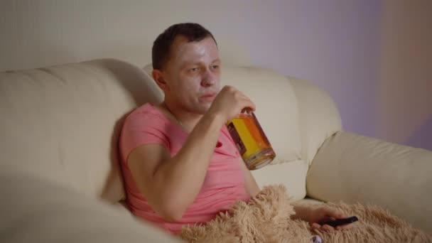 Un hombre con una máscara cosmética en la cara y una botella de alcohol en la mano mira la televisión por la noche - Imágenes, Vídeo