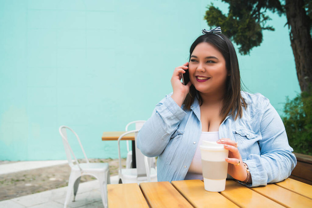 Portrét mladé plus velikost žena mluví do telefonu, zatímco sedí venku v kavárně. - Fotografie, Obrázek