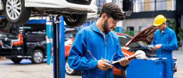 Kék munkaruhás kaukázusi szerelő egyenruhában ellenőrzi a jármű karbantartási ellenőrzőlistáját elmosódott autóval a háttérben. Gépjármű-javítási szolgáltatás, Szakmai foglalkozás. - Fotó, kép