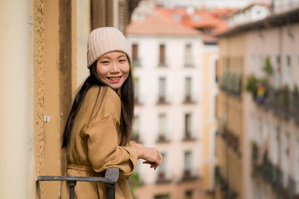 young happy and beautiful Asian Chinese woman in winter hat enjoying city view from hotel room balcony in Spain during holidays trip in Europe smiling cheerful in urban background - Фото, зображення