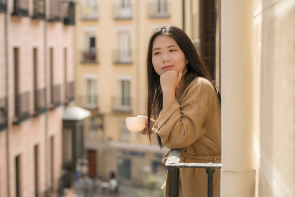 ヨーロッパ旅行中にスペインのホテルのバルコニーから街の景色を楽しむ若い幸せと美しいアジア系日本人女性都市部を背景に明るい笑顔コーヒーを飲む - 写真・画像